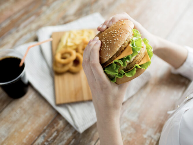 person holding fast food burger with onion rings and a soda on the table