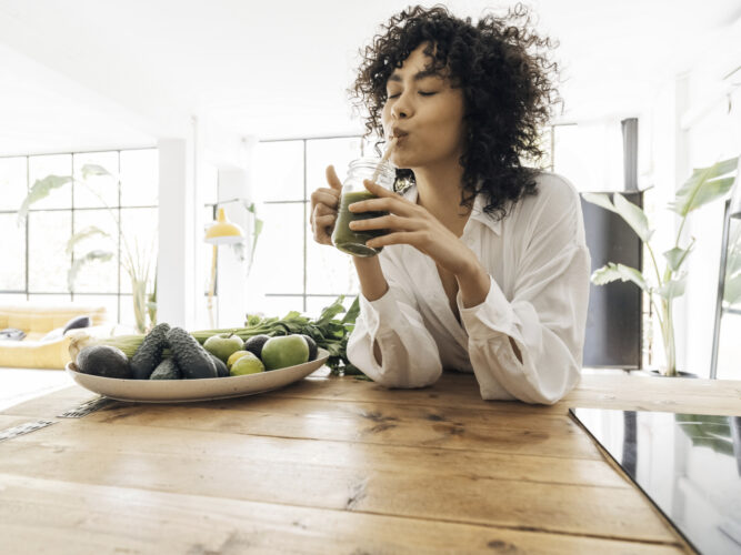 woman drinking green smoothie in a bright kitchen with healthy foods