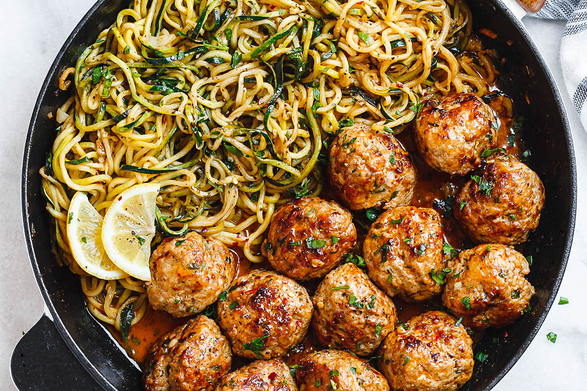 turkey meatballs in a skillet with cooked zoodles and lemon wedges